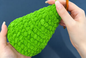 A plain green chenille cone held on its side in two hands against a dark blue background, its wide end open, with a gold crochet hook in the right hand
