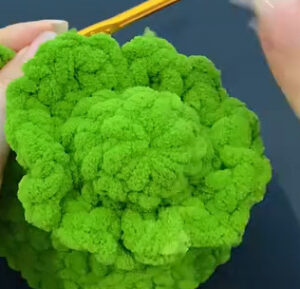The top of a green chenille tree seen from above, ruffled frills round its tip, with a yellow crochet hook at the edge of the ruffle and fingers holding the piece