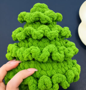 A green chenille tree with four tiers of ruffled frills and no pompoms yet, steadied at its base by two fingers, against a dark background