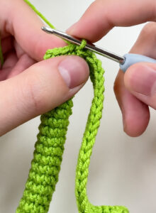 The top of a green crocheted leg held between finger and thumb, a long crocheted chain looping down from it, and a crochet hook in the stitches at the top