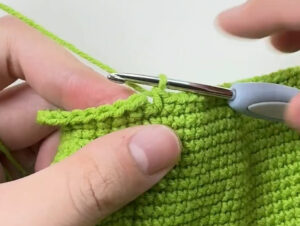 The top edge of a green crocheted piece held flat under a thumb, a crocheted chain lying along the edge and a crochet hook in the stitch at the end of it