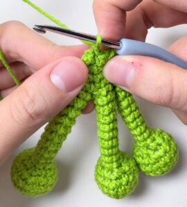 Three green crocheted toes, each a small ball on a narrow tube, held side by side between finger and thumb, with a crochet hook in the stitches that join them at the top